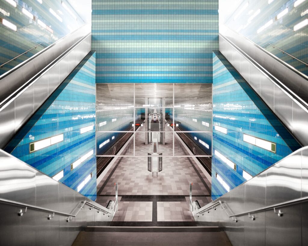 Symmetrische Ansicht einer modernen, leeren U-Bahn-Station mit blau gekachelten Wänden, Metallgeländern und zwei Rolltreppen, die zum darunter liegenden Bahnsteig hinunterführen.