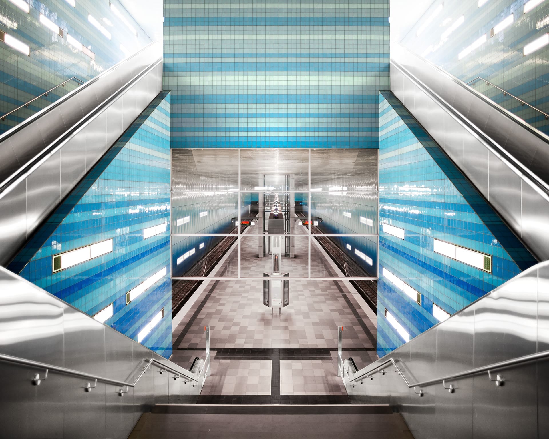 Symmetrische Ansicht einer modernen, leeren U-Bahn-Station mit blau gekachelten Wänden, Metallgeländern und zwei Rolltreppen, die zum darunter liegenden Bahnsteig hinunterführen.