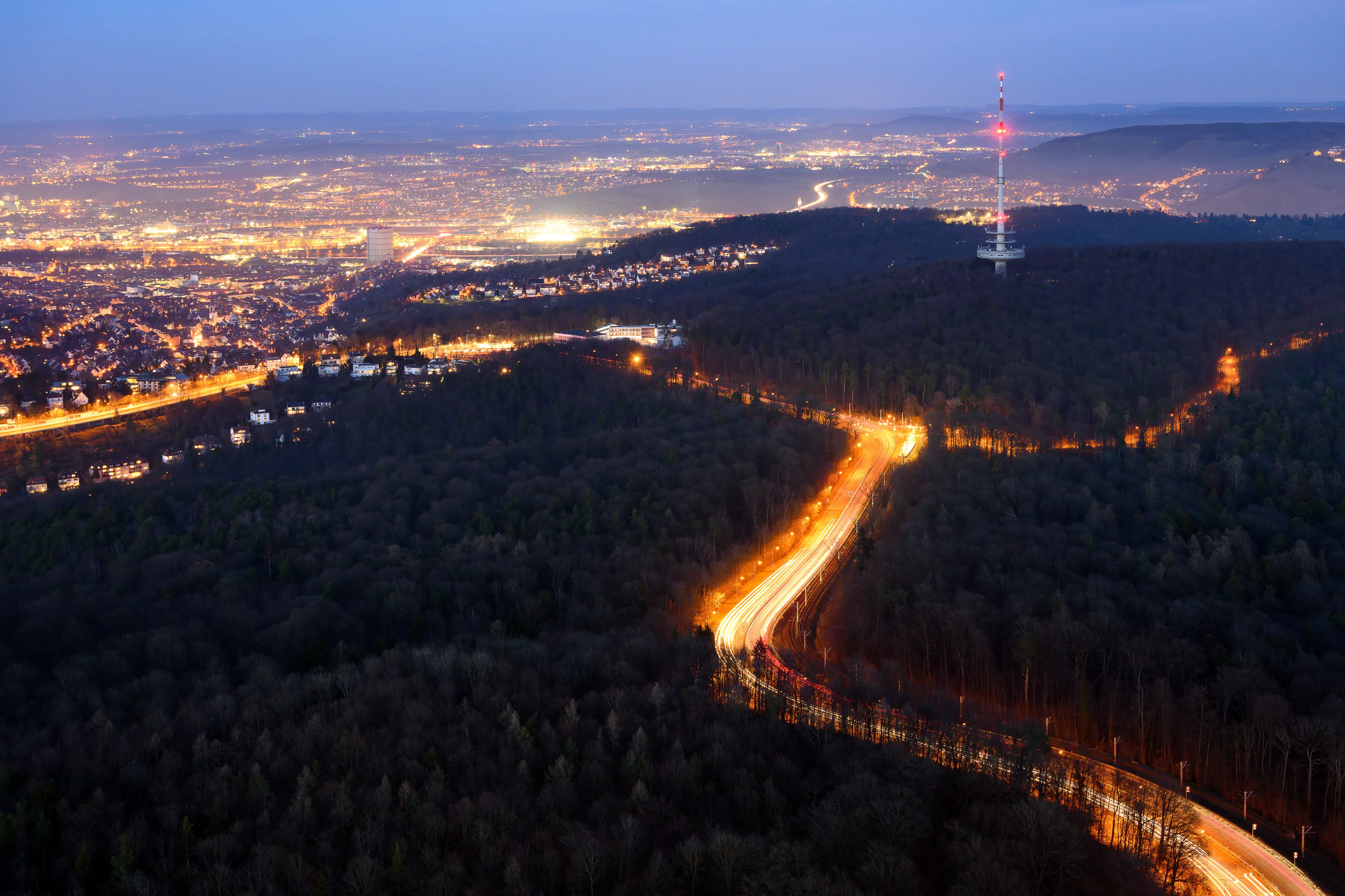 Luftaufnahme einer Stadt in der Abenddämmerung mit einer Autobahn, die sich durch einen Wald schlängelt, sichtbaren Stadtlichtern und einem großen Funkturm auf einem Hügel.