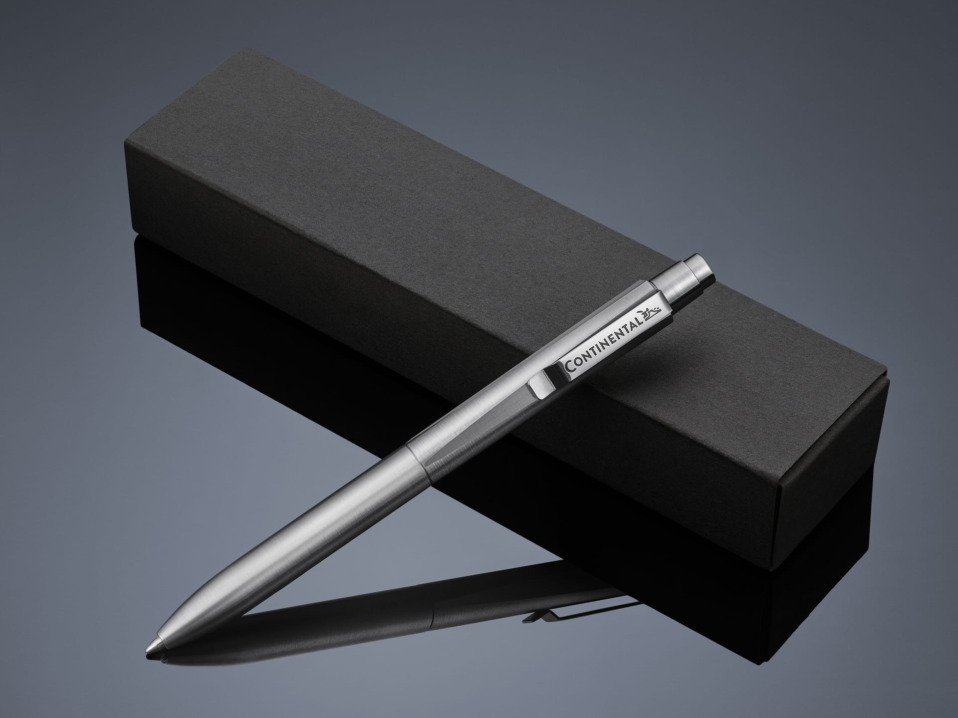 Ein Metallkugelschreiber mit dem Schriftzug CONTINENTAL" ruht diagonal auf einem geschlossenen schwarzen, rechteckigen Kasten, der sich von einer dunklen, reflektierenden Oberfläche abhebt.