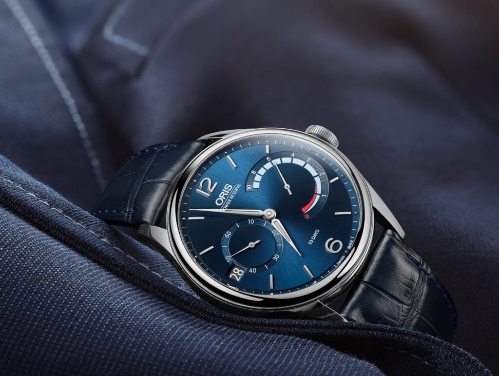 Nahaufnahme einer blauen Oris-Armbanduhr mit Lederband, Ziffernblättern und Gangreserveanzeige, die auf einer marineblauen Stoffunterlage ruht.