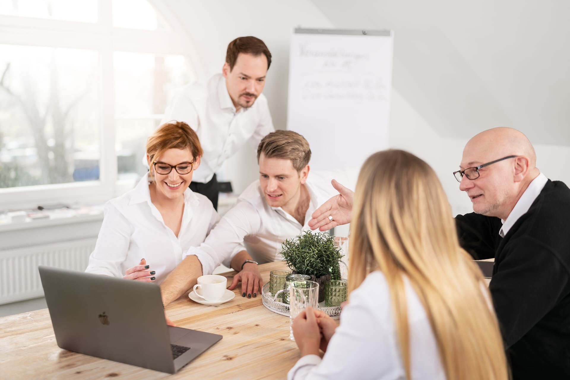 Fünf Personen sitzen um einen Tisch herum, schauen auf einen Laptop und diskutieren in einer hellen Büroumgebung gemeinsam über etwas.