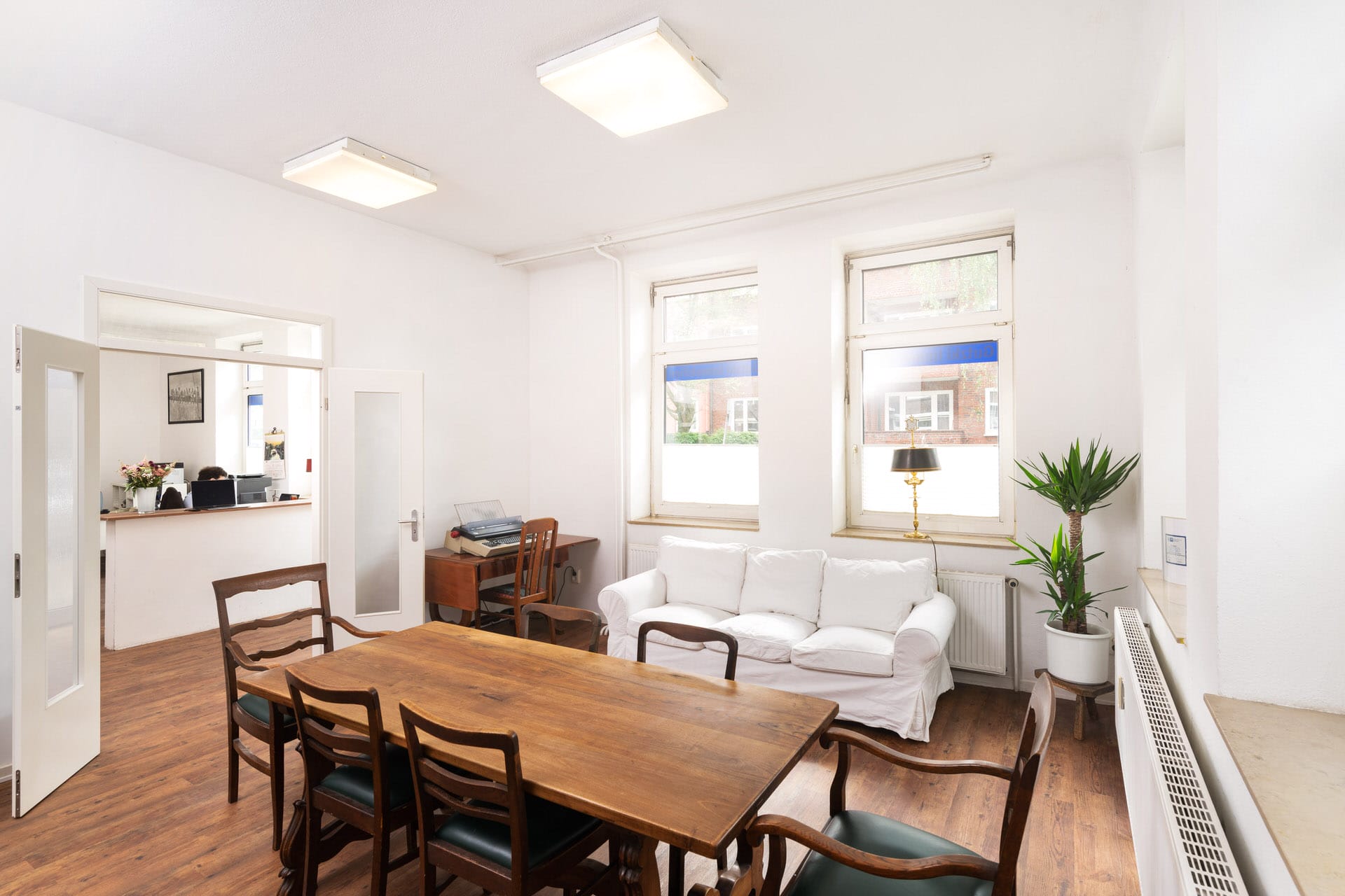 Heller Büroraum mit einem Holztisch, sechs Stühlen, einem weißen Sofa, Zimmerpflanzen, einer kleinen Lampe und großen Fenstern, die natürliches Licht hereinlassen.