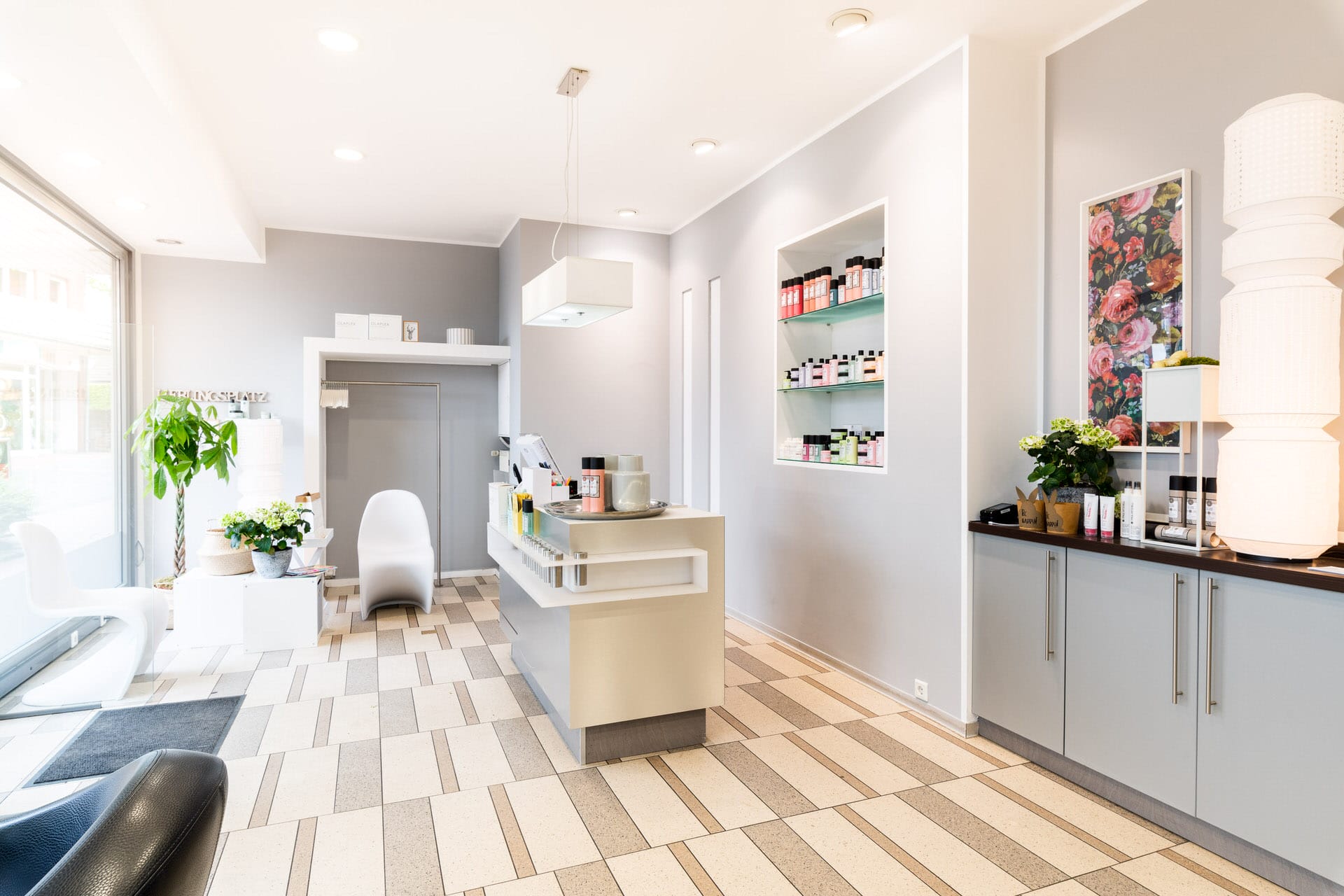 Moderner, heller Salon mit Empfangstresen, Produktregalen, Stühlen, Topfpflanzen und einem großen Fenster, das natürliches Licht hereinlässt.