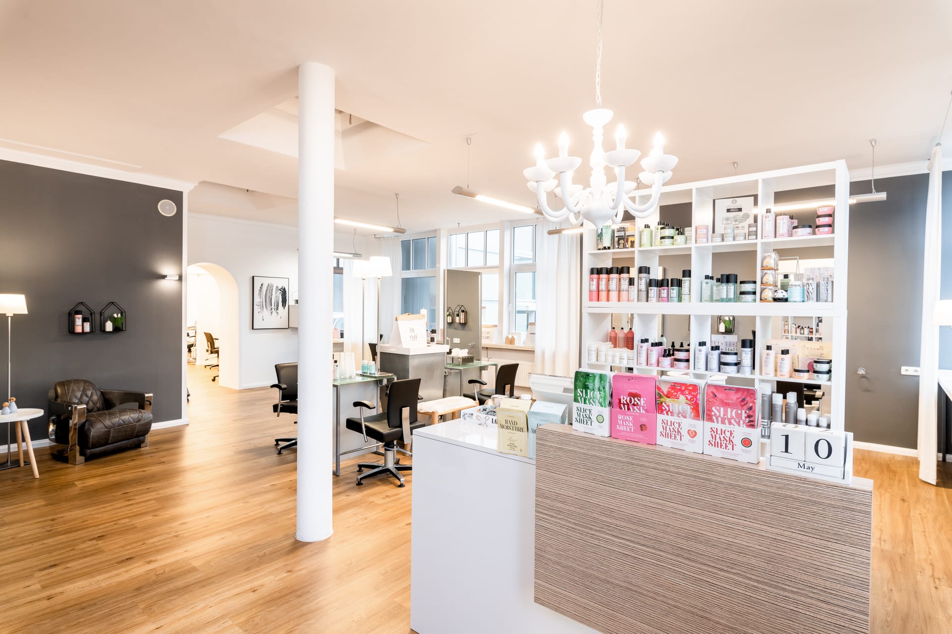 Ein moderner, heller Salon mit Holzböden, Stylingstationen, Produktregalen, einem Empfangstresen und einem weißen Kronleuchter.
