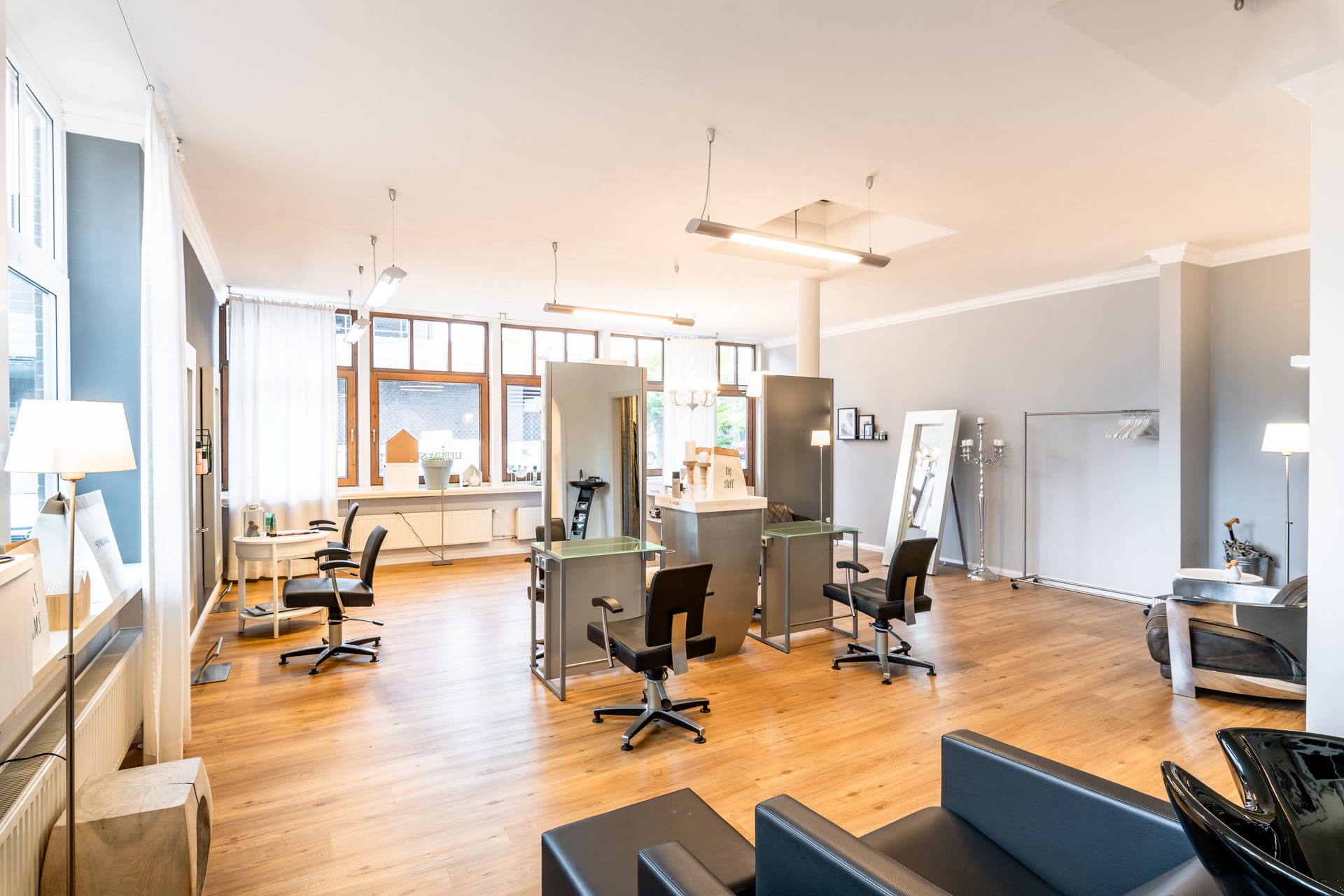 Moderner Friseursalon mit Frisierplätzen, Spiegeln, schwarzen Stühlen, großen Fenstern, Holzboden und neutralen Wänden.