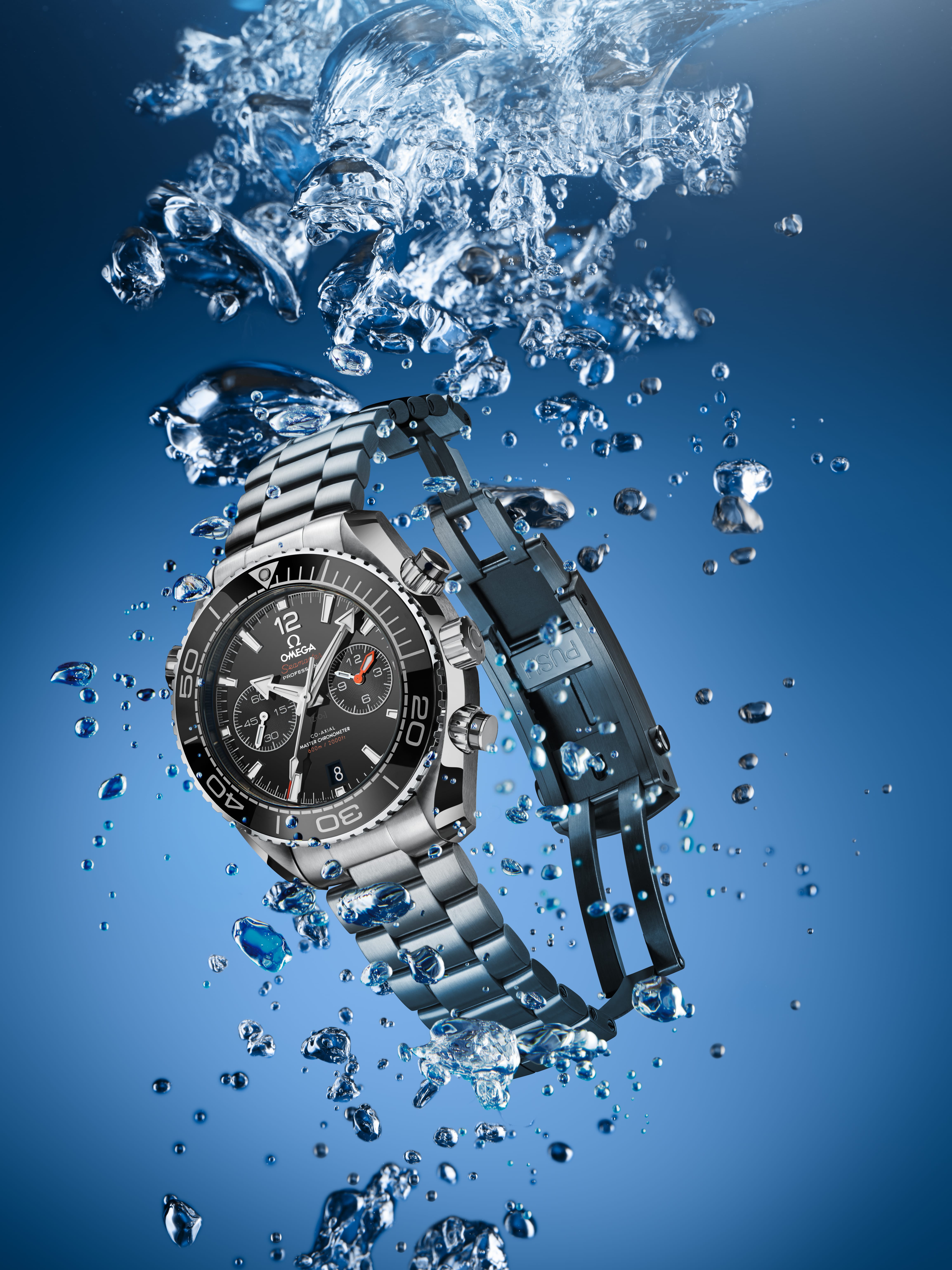 Eine silberne Armbanduhr mit schwarzem Zifferblatt ist in Wasser getaucht, umgeben von spritzenden Tropfen vor einem blauen Hintergrund.