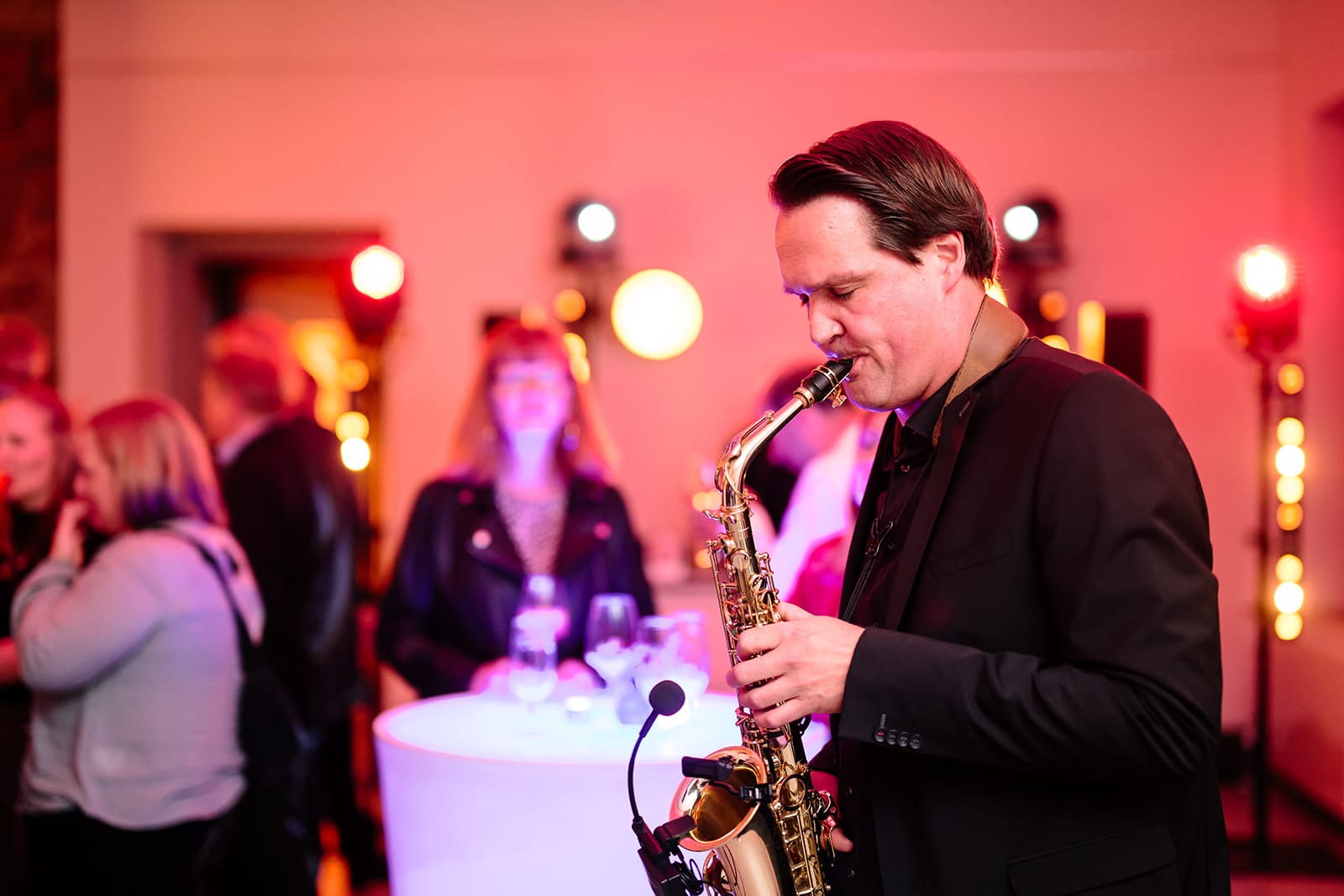 Ein Mann in einem schwarzen Anzug spielt Saxophon bei einer Veranstaltung in einer Halle, während sich im Hintergrund Leute treffen.