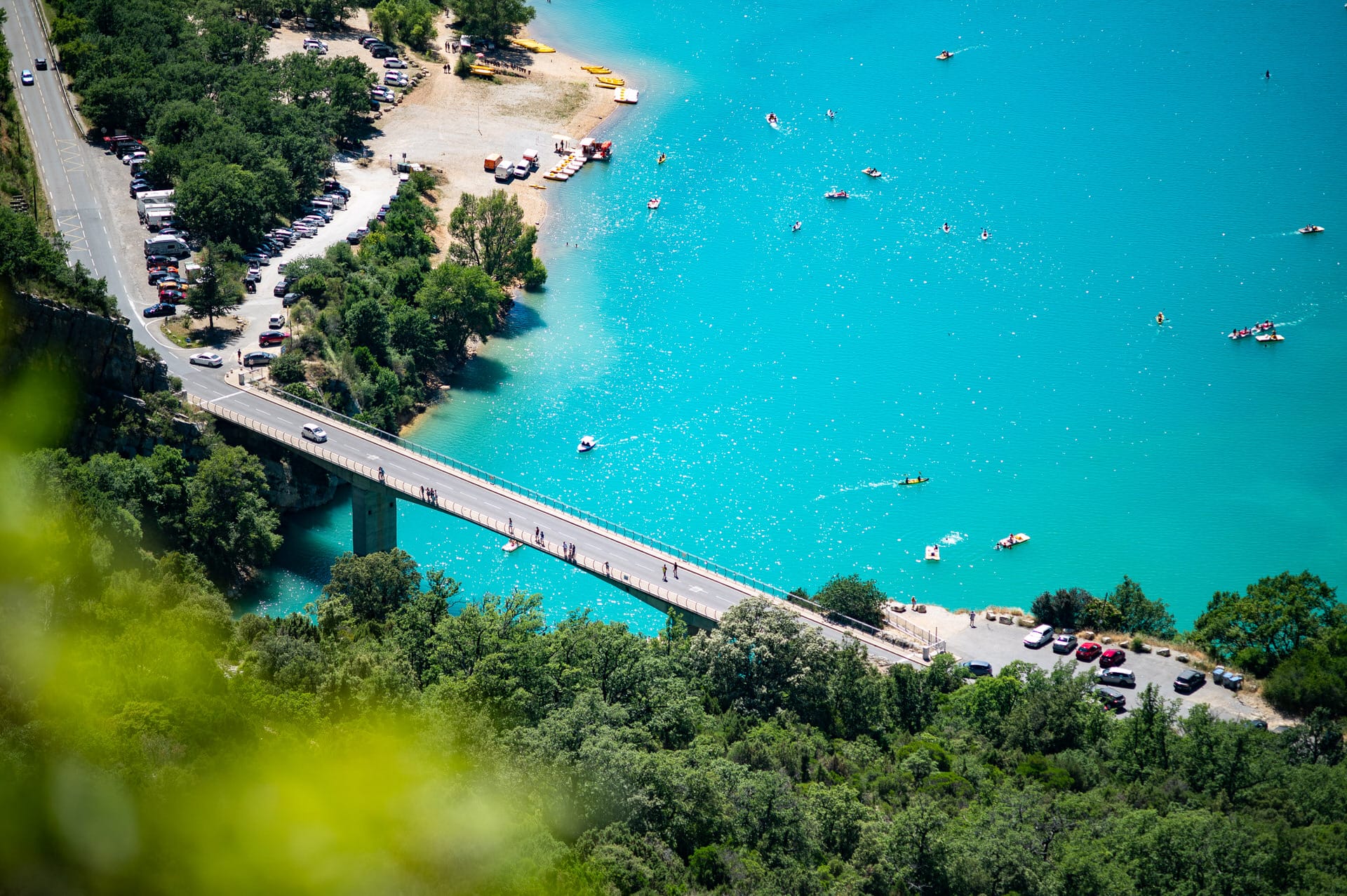 Eine Brücke führt über hellblaues Wasser, in deren Nähe Autos geparkt sind; man sieht Menschen auf der Brücke und in Booten auf dem Wasser, umgeben von dichten grünen Bäumen.