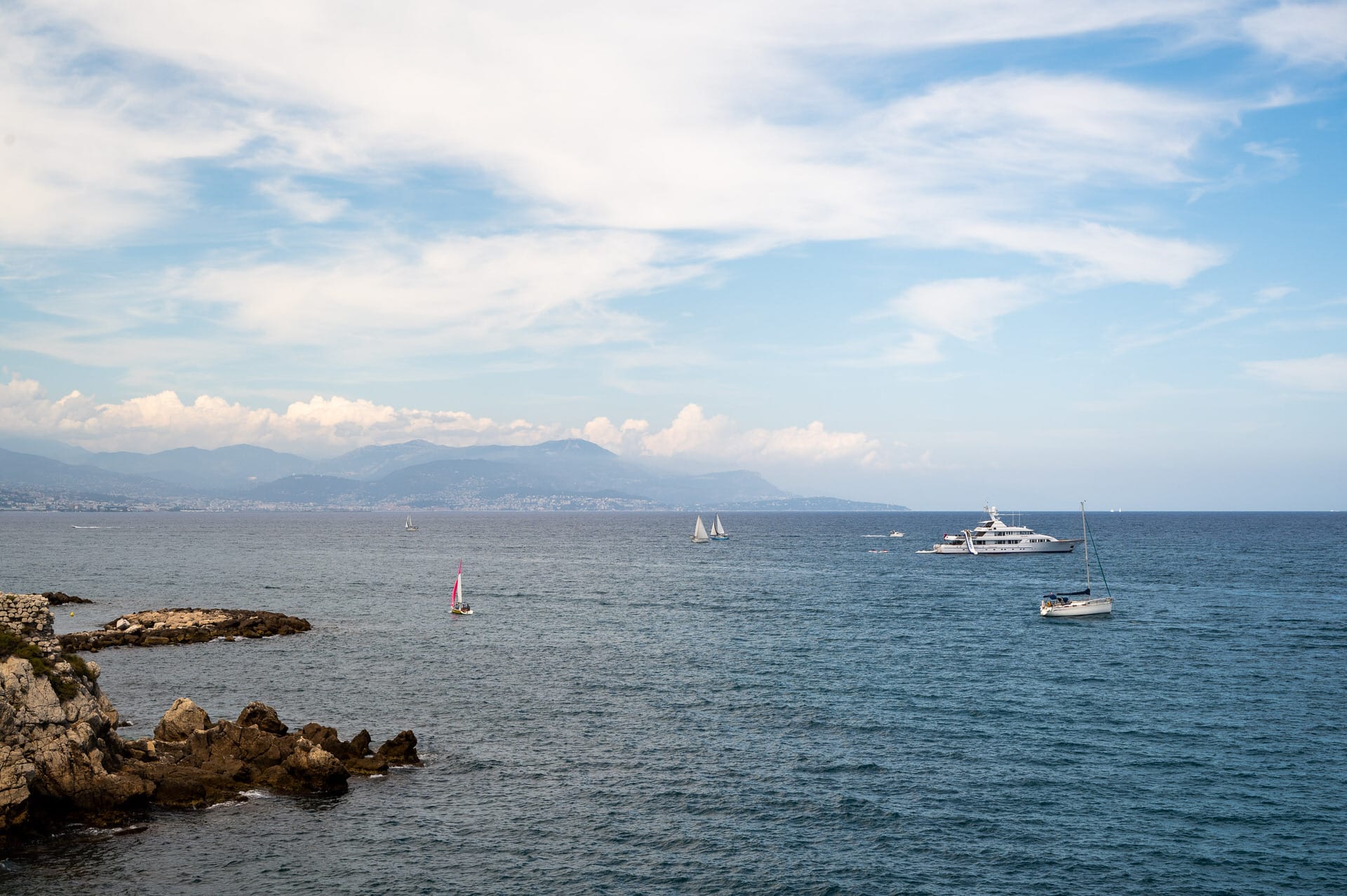 Küstenszene mit felsiger Küste auf der linken Seite, mehreren Segelbooten und einer Yacht auf dem Wasser und entfernten Bergen unter einem teilweise bewölkten Himmel.