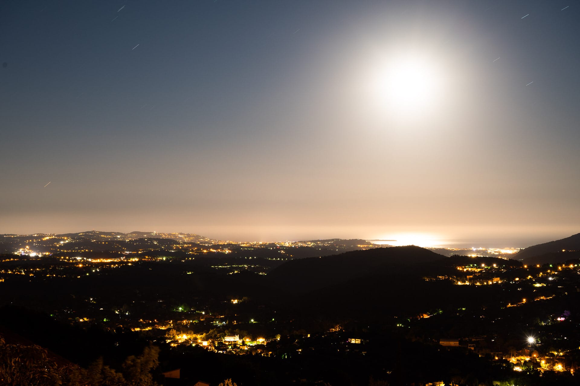Eine nächtliche Landschaft zeigt einen hell erleuchteten Himmel mit einem großen Mond, die Lichter der Stadt, die über die Hügel verstreut sind, und Spiegelungen auf einem entfernten Gewässer.