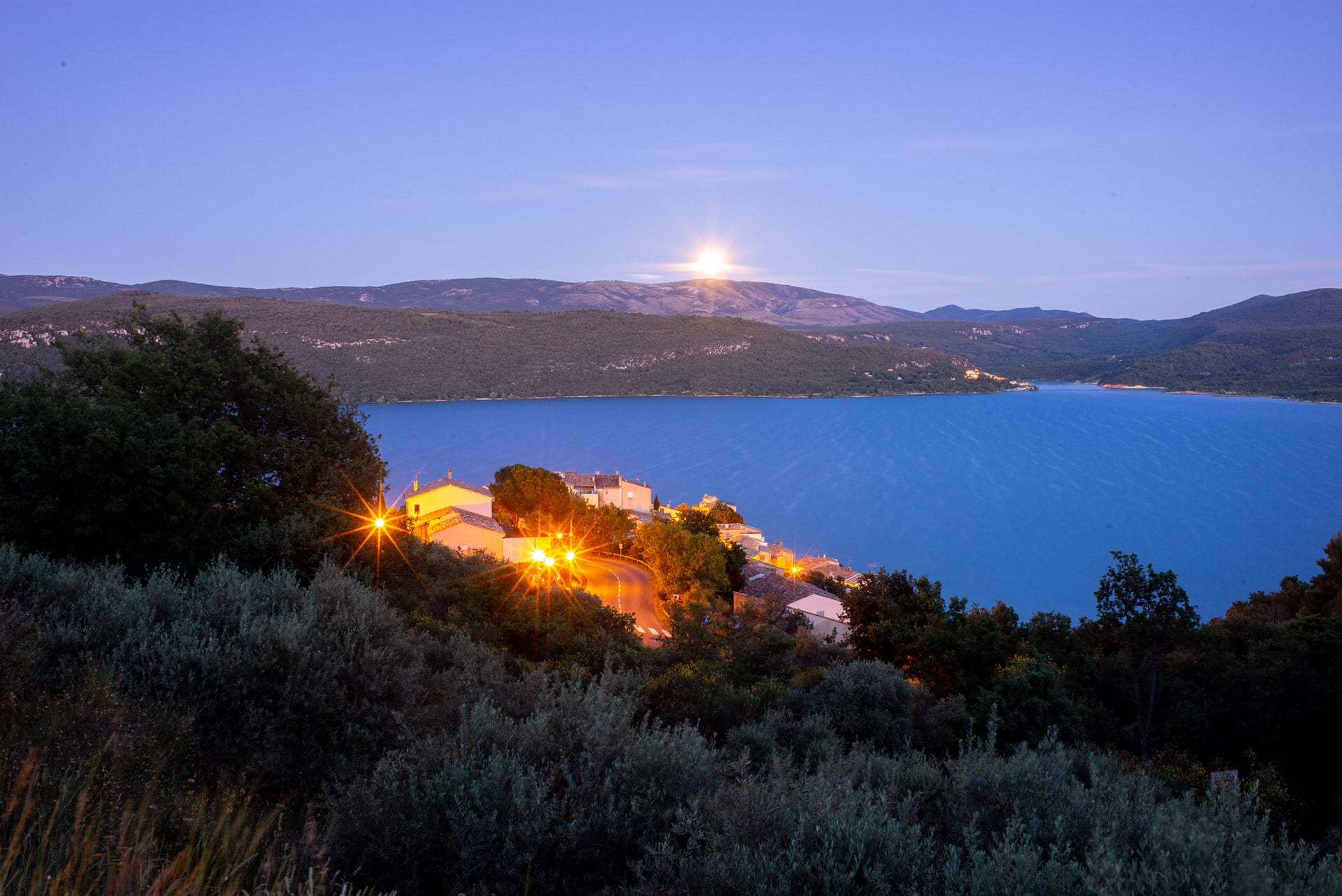 Ein Küstendorf mit beleuchteten Straßenlaternen liegt an einem blauen Gewässer, umgeben von Hügeln, während der Mond in der Abenddämmerung über dem Horizont aufgeht.