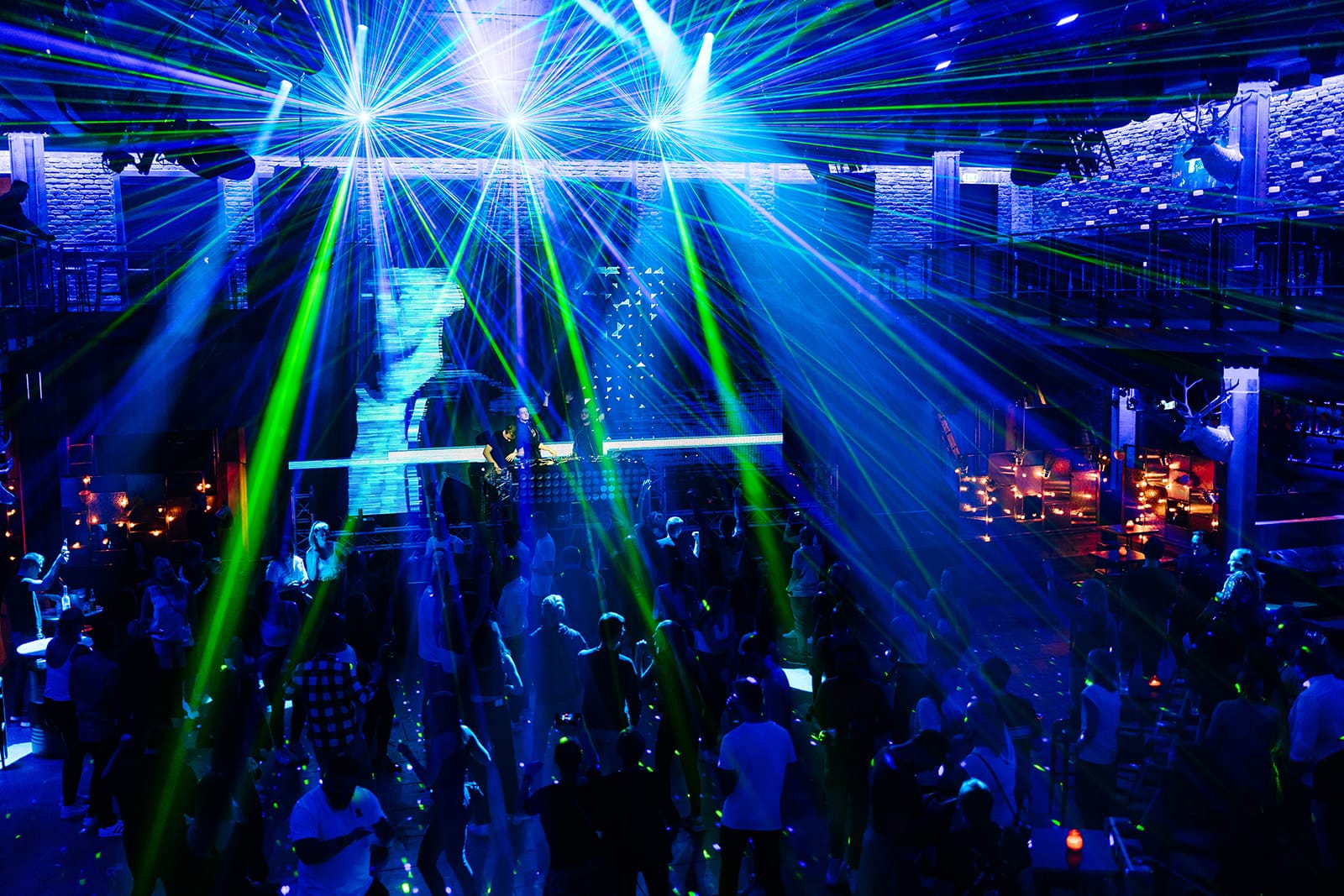 Eine überfüllte Tanzfläche in einem Nachtclub mit tanzenden Menschen unter blauem und grünem Laserlicht, umgeben von beleuchteten Tischen und einem Balkon.