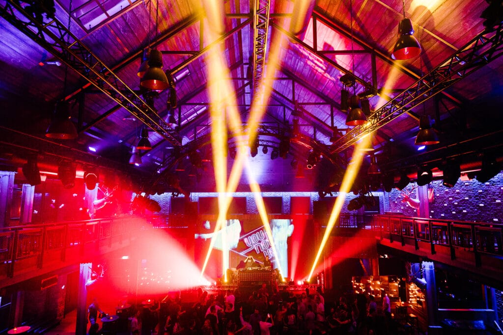 Großer überdachter Nachtclub mit bunten Bühnenlichtern, einer hohen Decke, einem erhöhten DJ-Pult und auf dem Boden tanzenden Menschen.