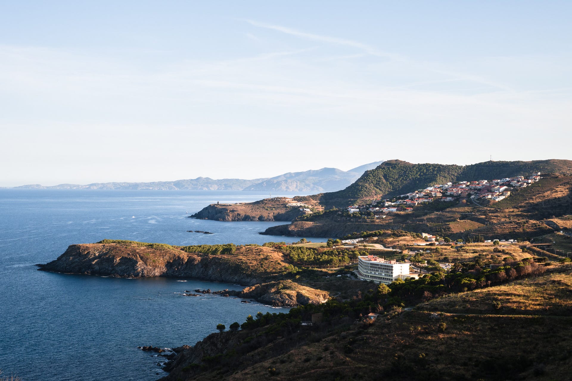 Eine Küstenlandschaft mit felsigen Halbinseln, einem kleinen weißen Gebäude in Küstennähe und einer Ansammlung von Häusern auf einem Hügel unter klarem Himmel. In der Ferne sind Berge zu sehen.