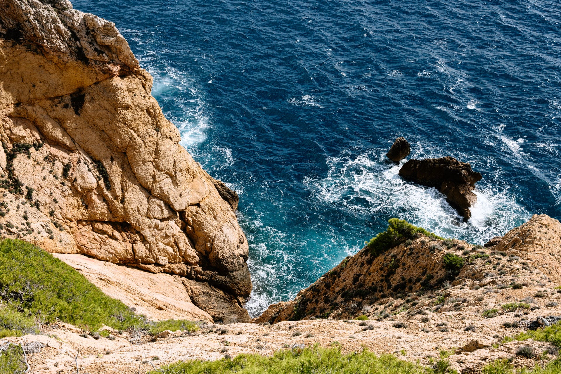 Steile Felsklippen mit spärlicher Vegetation und Blick auf den tiefblauen Ozean, wo die Wellen gegen die Felsen schlagen.