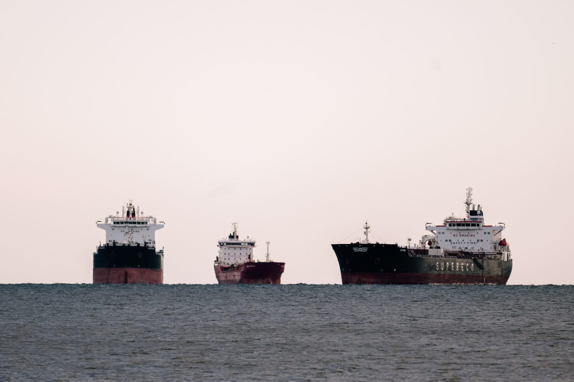 Drei große Frachtschiffe liegen im Meer vor Anker, aufgereiht vor einem blassen Himmel.