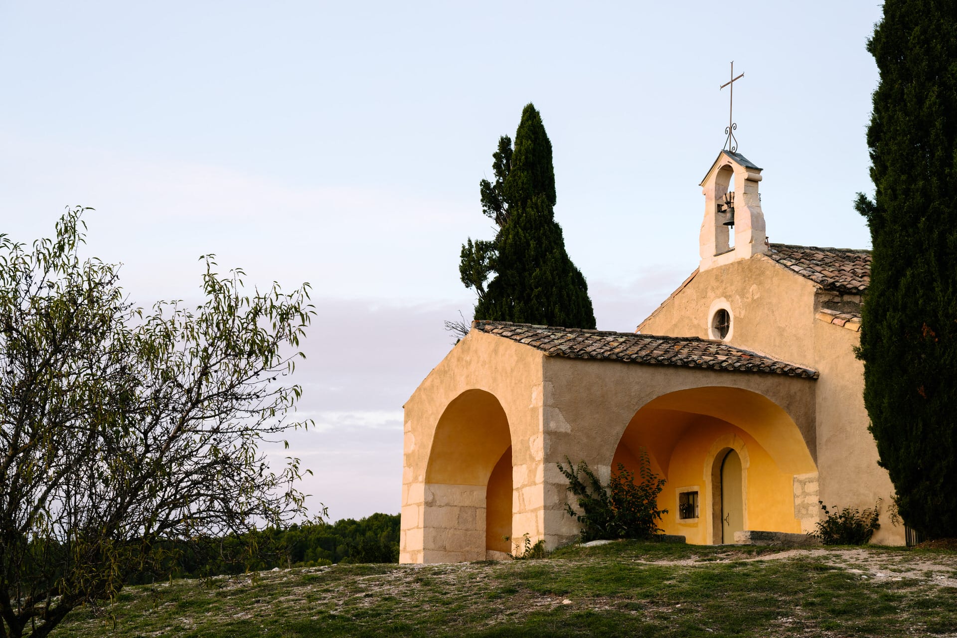 Kleine, rustikale Kapelle mit gewölbtem Eingang, Ziegeldach und einem Glockenturm, umgeben von Bäumen und Grün unter einem klaren Himmel.