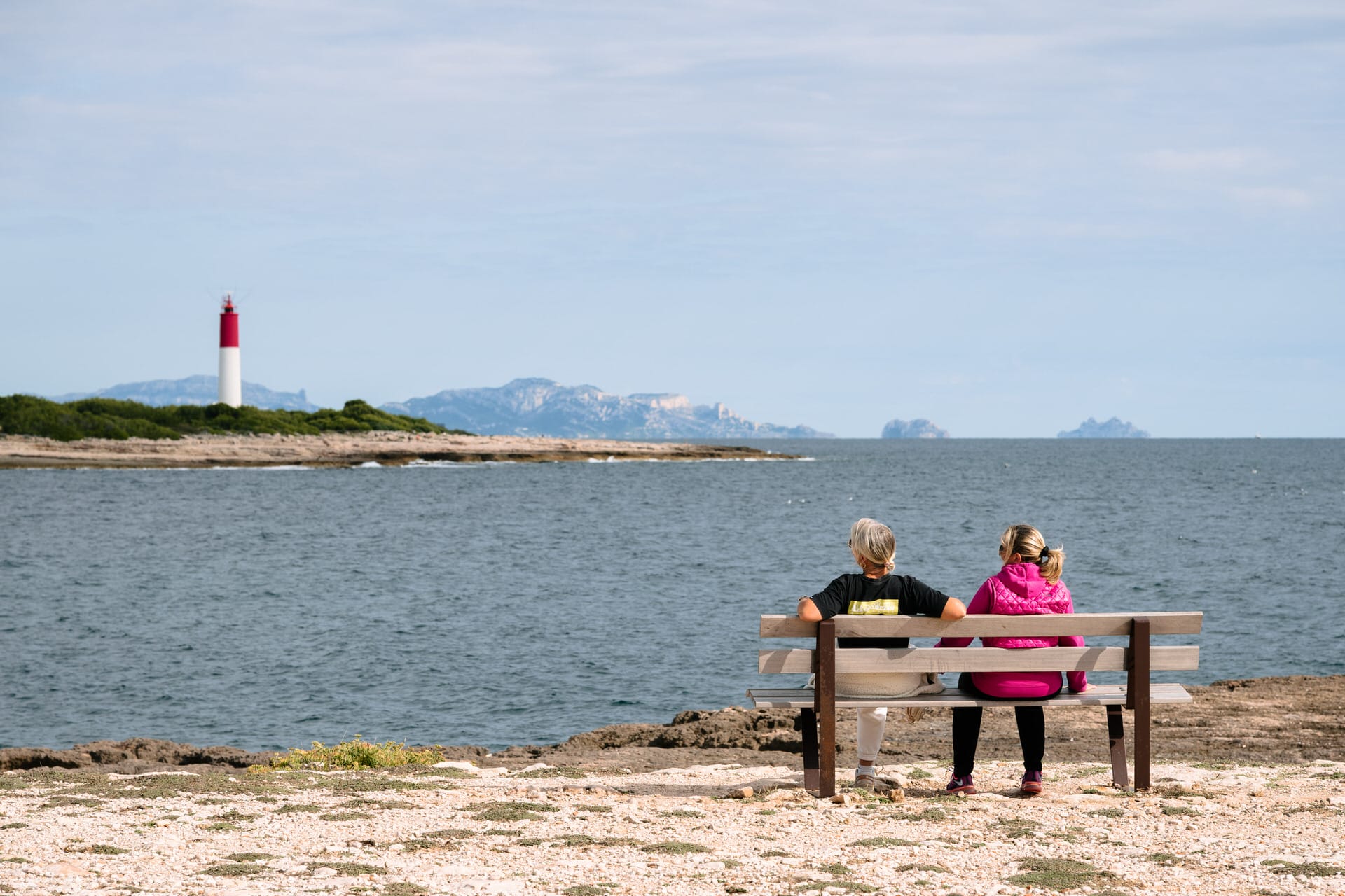 Zwei Personen sitzen auf einer Bank am Ufer und blicken auf das Meer, während in der Ferne ein rot-weißer Leuchtturm und die Berge im Hintergrund zu sehen sind.