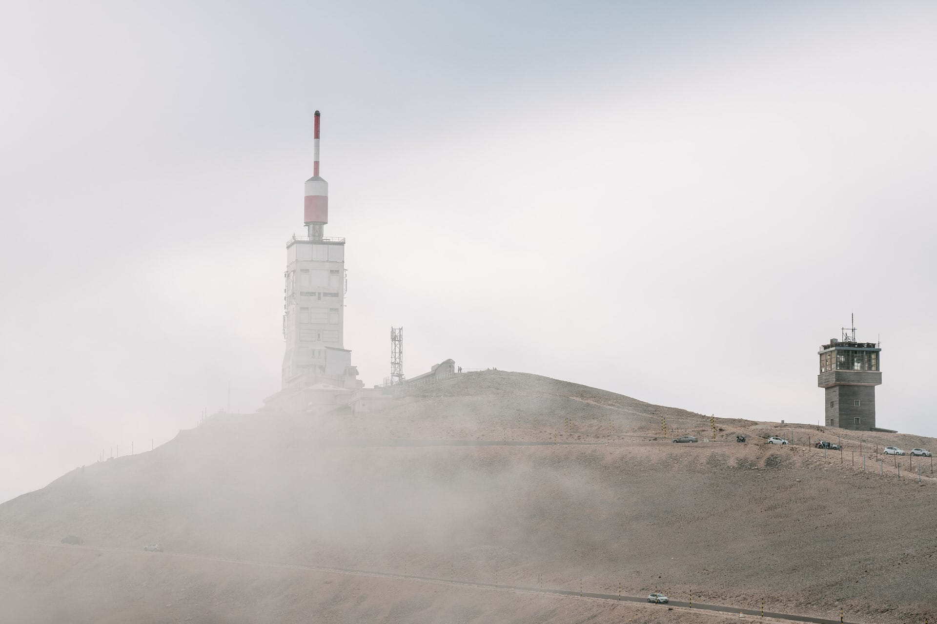 Ein hoher weißer Turm mit einer rot gestreiften Antenne steht auf einem nebligen, felsigen Hügel, in dessen Nähe sich ein kleineres Gebäude und mehrere geparkte Autos befinden.