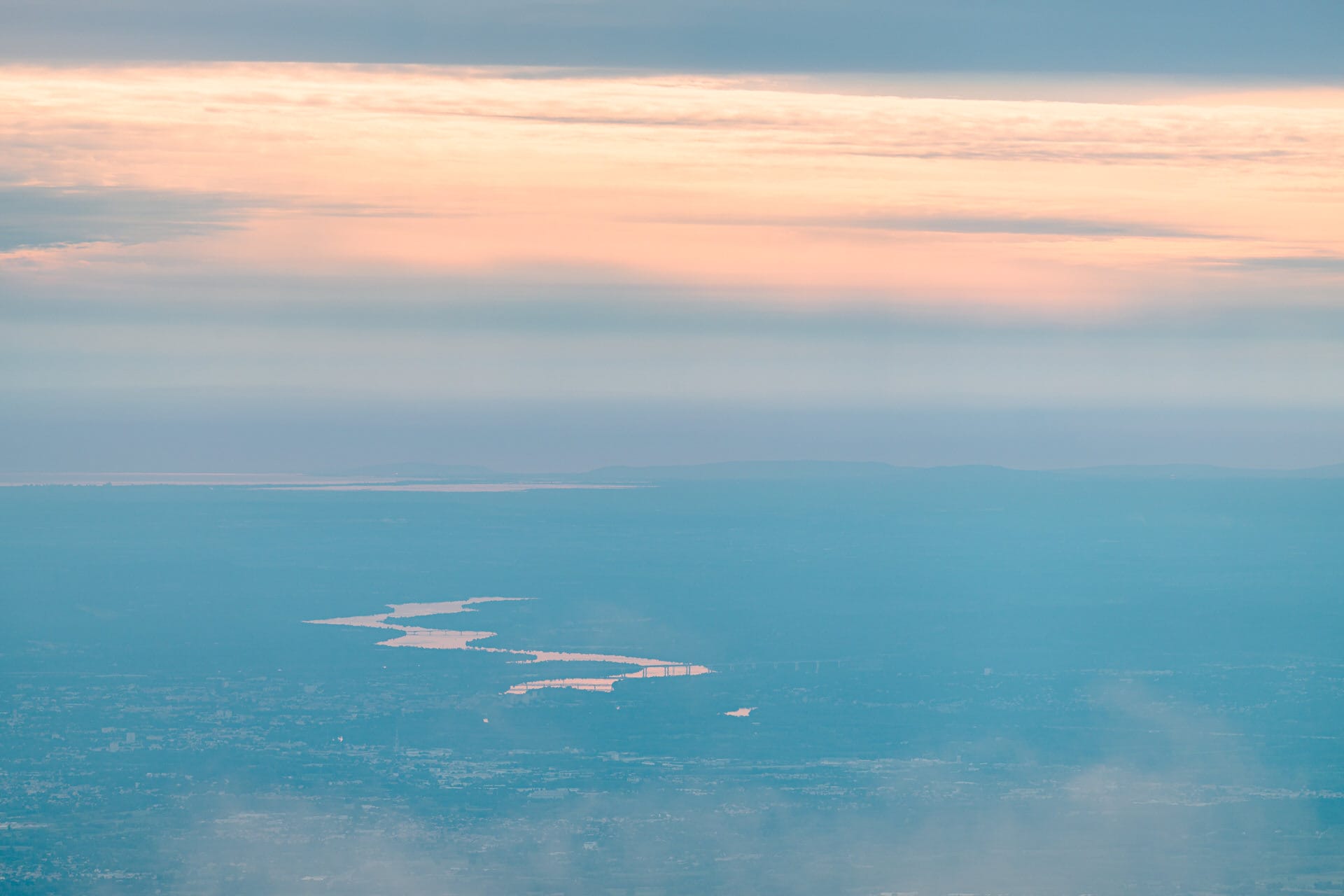 Luftaufnahme eines sich schlängelnden Flusses, in dem sich das Licht unter einem pastellfarbenen Himmel mit vereinzelten Wolken und entferntem Land am Horizont spiegelt.