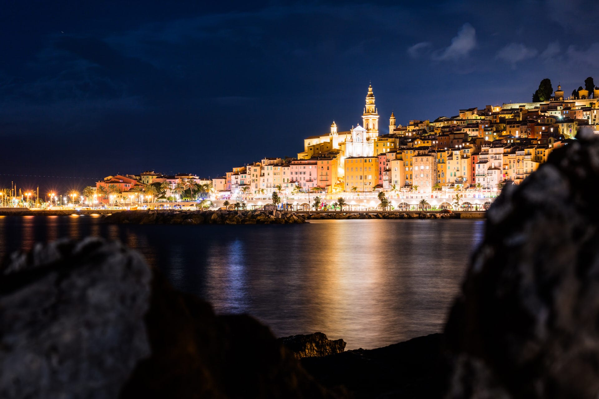 Nächtliche Ansicht einer Küstenstadt mit beleuchteten Gebäuden, die sich auf dem Wasser spiegeln, dunklen Felsen im Vordergrund und einem Kirchturm, der sich von den anderen Gebäuden abhebt.
