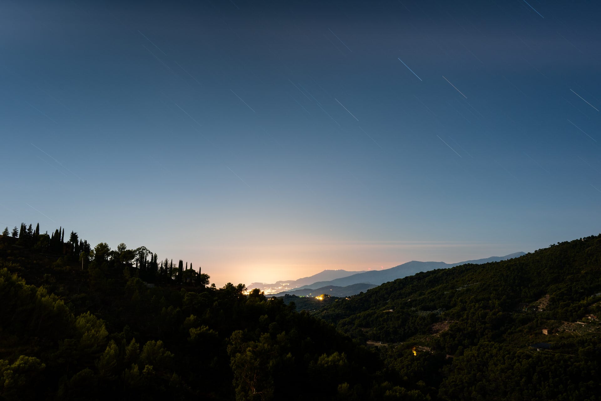 Nächtliche Landschaft mit Sternenspuren am Himmel, baumbewachsenen Hügeln im Vordergrund und entfernten Stadtlichtern, die in der Nähe des Horizonts leuchten.