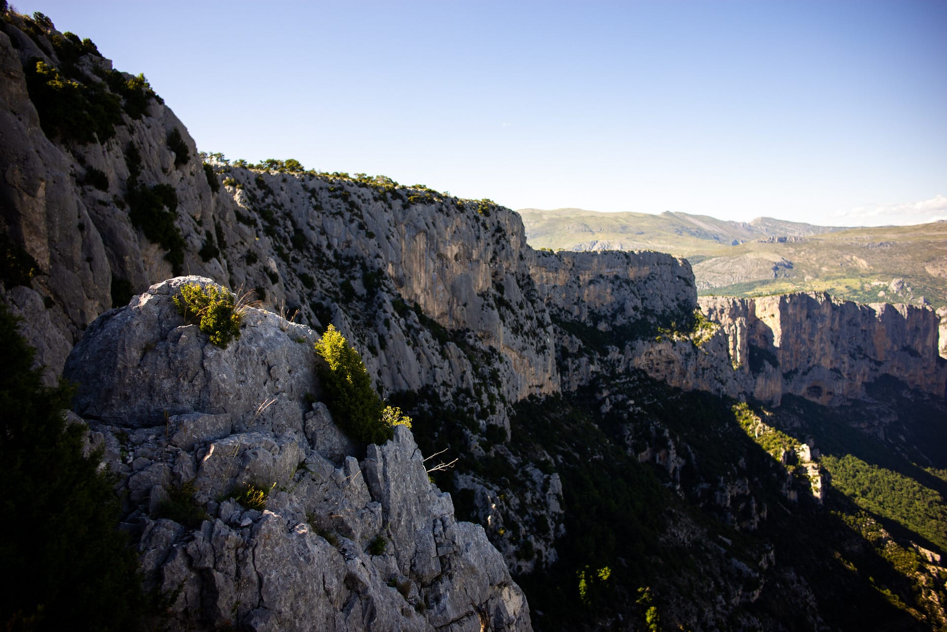 Ein felsiger Abhang mit spärlicher Vegetation, der ein tiefes Tal überblickt, unter einem klaren blauen Himmel, mit fernen Hügeln im Hintergrund.