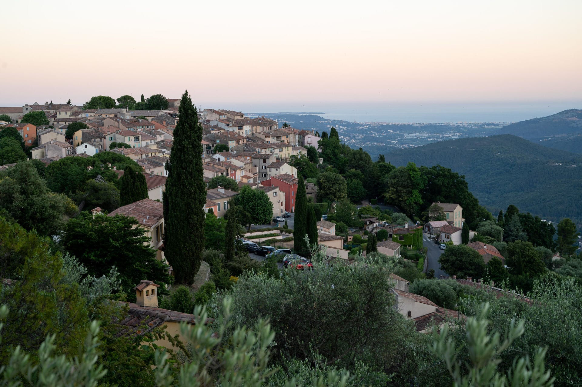 Ein Dorf am Hang mit terrakottafarbenen Häusern, Zypressen und geparkten Autos überblickt die grünen Hügel und das ferne Meer bei Sonnenuntergang.