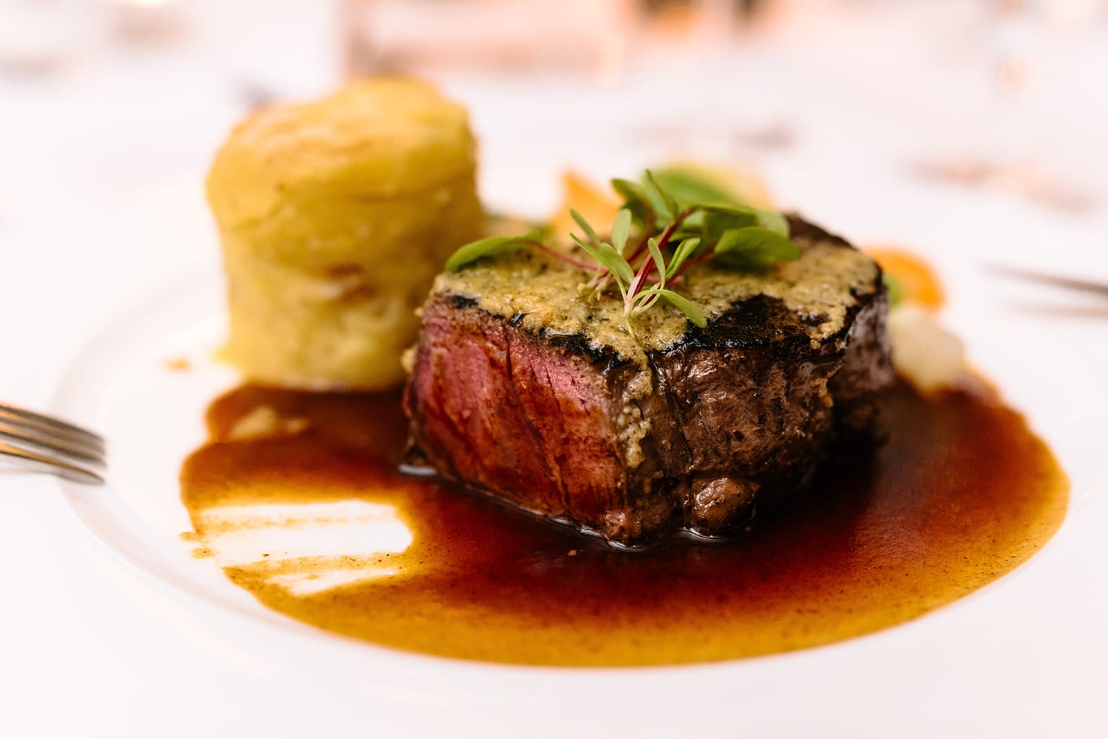 Ein Tellergericht mit einem medium-rare Steak mit Kräutertopping, brauner Soße, einer Portion geschichteter Kartoffeln und garniert mit Mikrogrün.