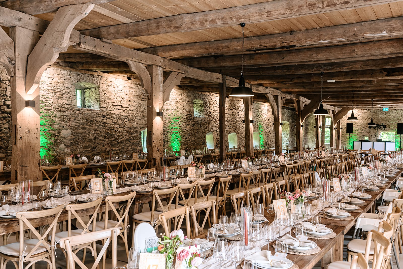 Ein rustikaler Bankettsaal mit langen Holztischen, die für eine Veranstaltung gedeckt sind, mit Steinwänden, Holzbalken und einfachen Blumensträußen.